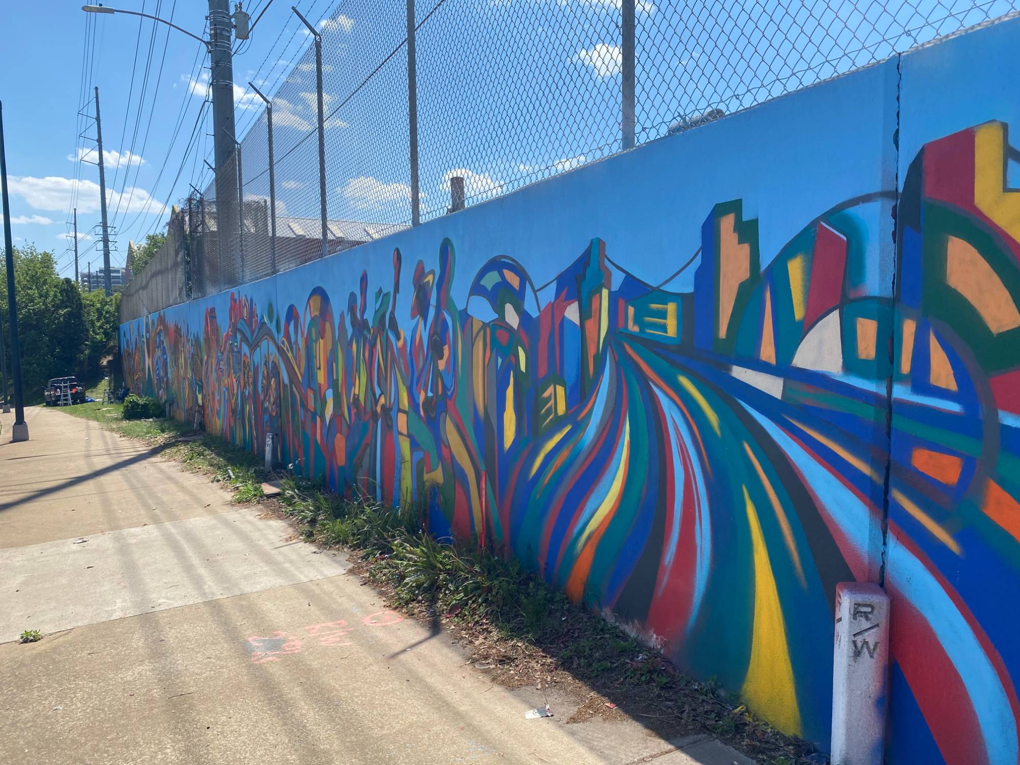 Long blue sidewalk wall mural with abstract figures, skyline shapes, and sweeping color streams