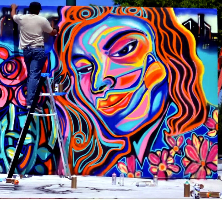 Muralist Corey Barksdale on a ladder spray painting a large portrait of a woman surrounded by flowers