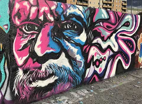 Street mural portrait of a bearded man in pink, black, and blue on a concrete wall